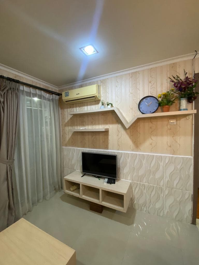 Ruang keluarga dengan televisi, AC terpasang, rak dinding, dan gorden di unit sewa Green Central City